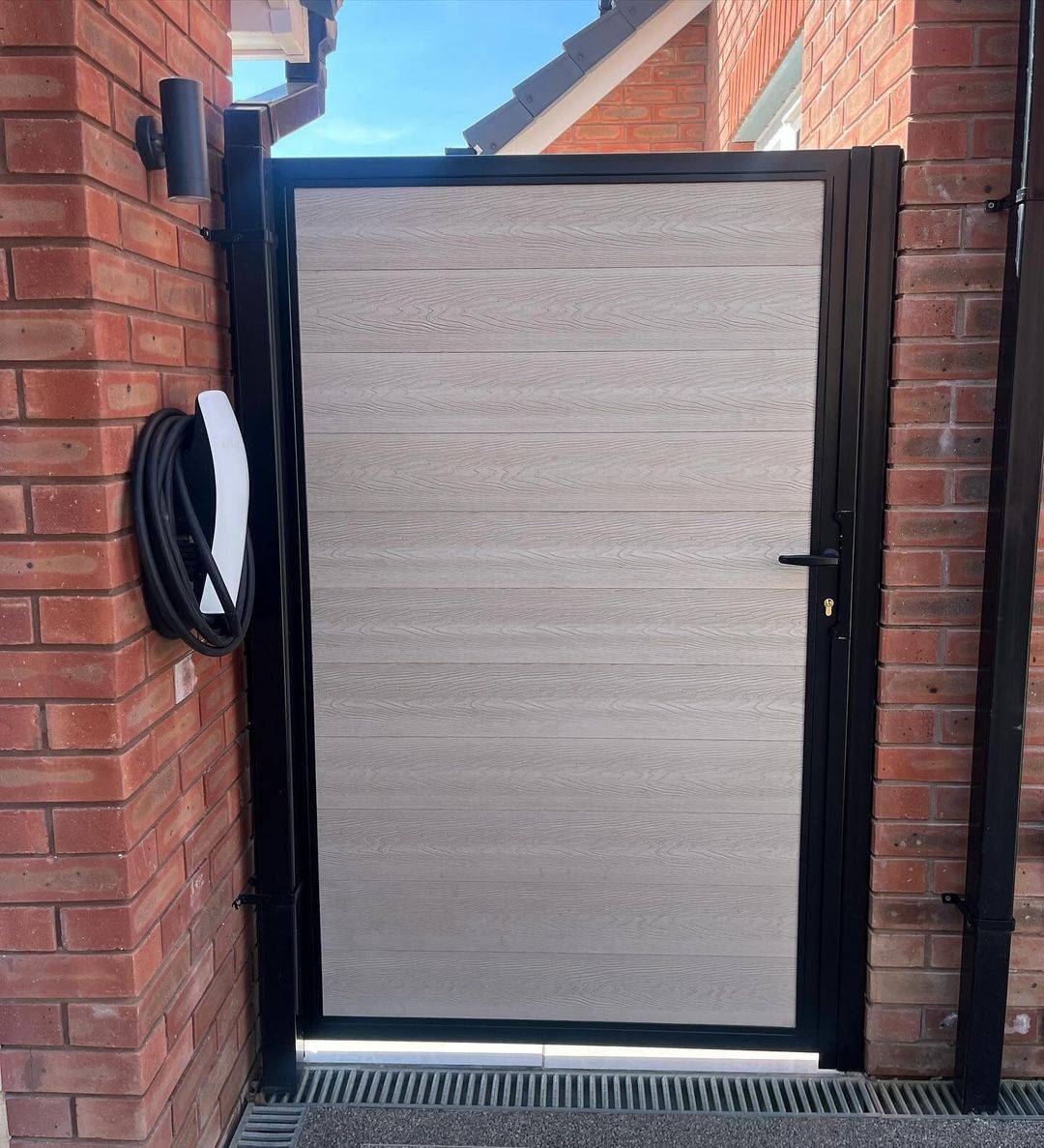 Solid composite wood-effect infill pedestrian gate in a black steel frame, mounted on a brick pier