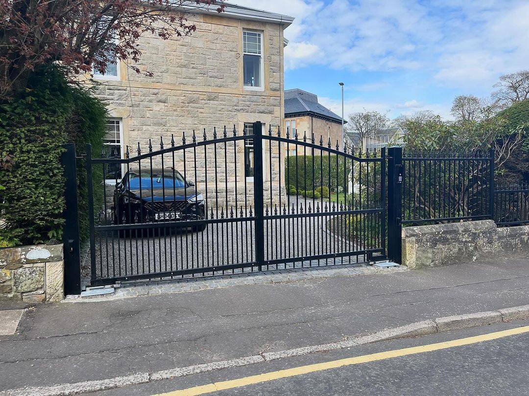 Simple black tubular steel arched-top double driveway gate with a stone building behind